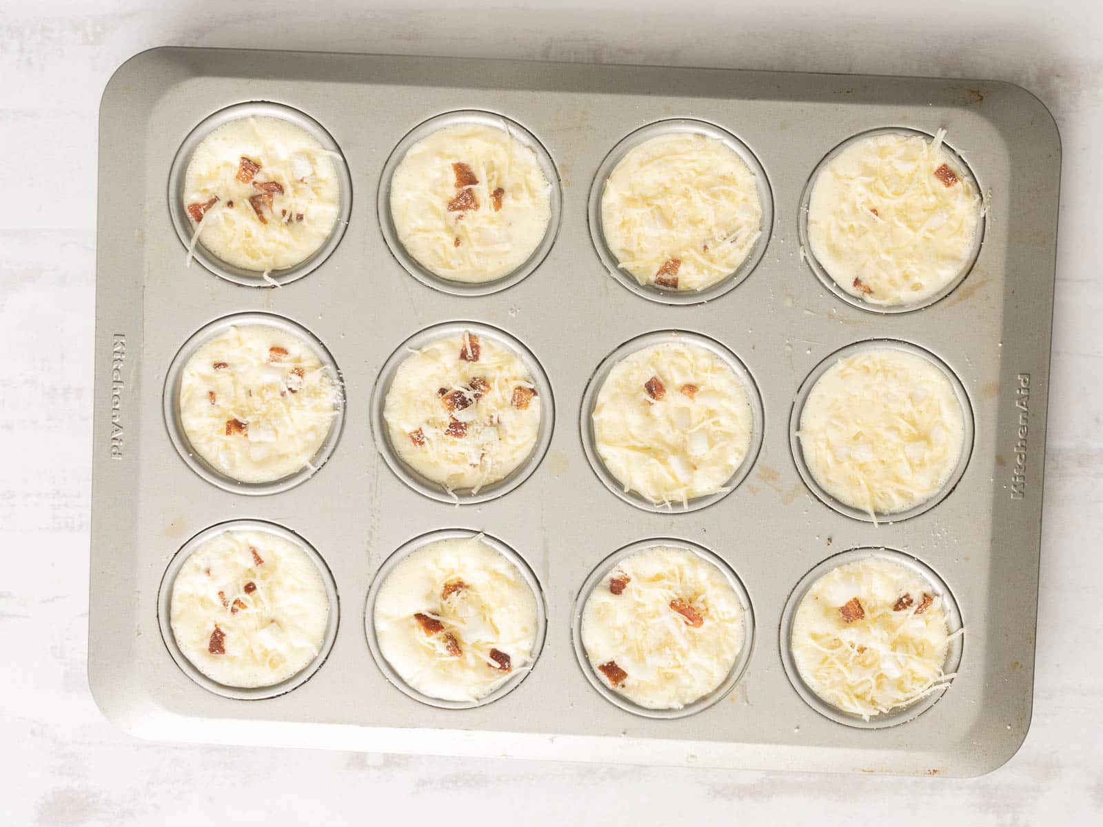 Quiche muffins filled with cheesy batter and bacon bits, viewed from above on a white surface.