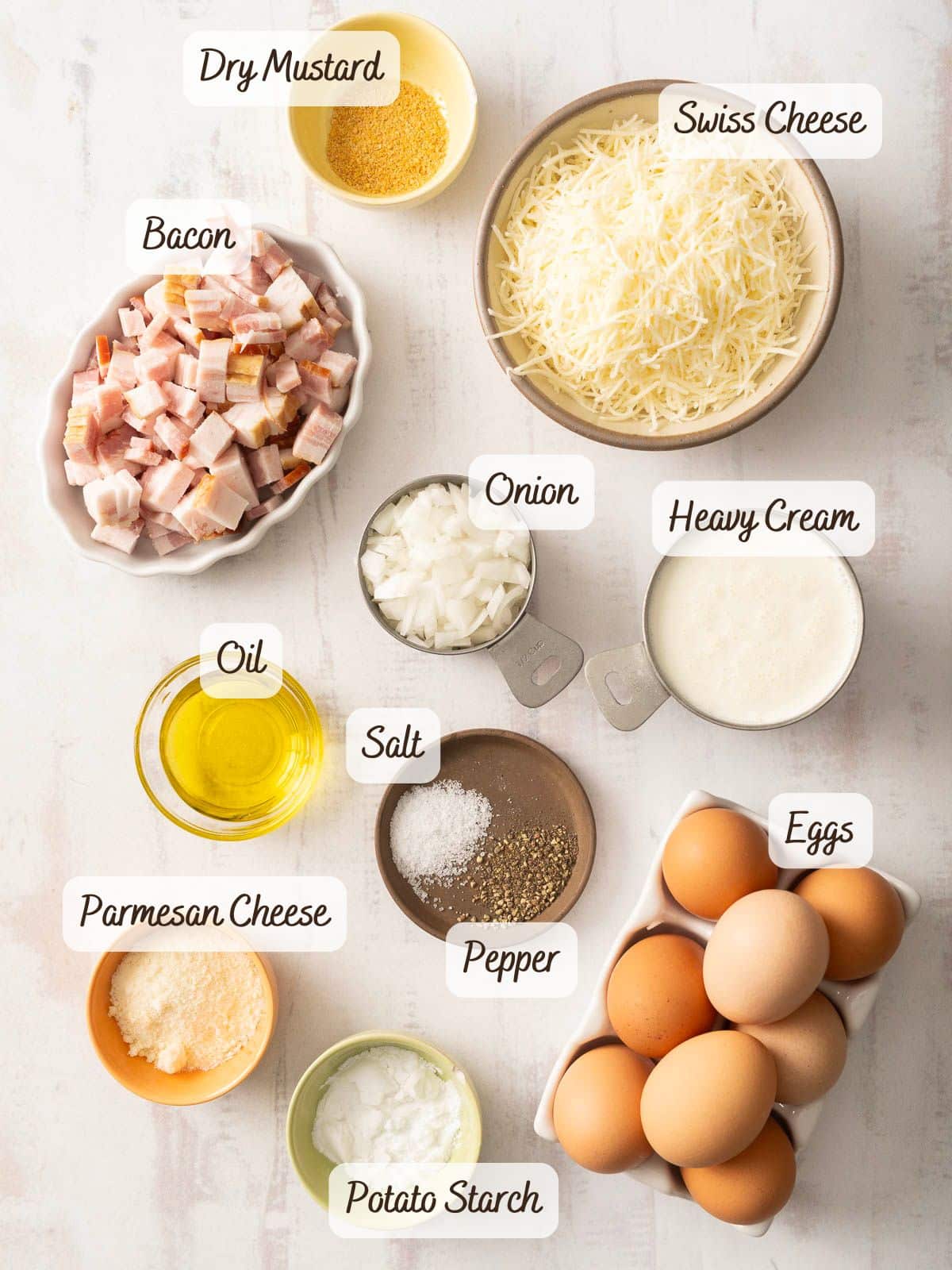 Ingredients arranged on a table for delectable quiche muffins, including swiss cheese, bacon, onion, heavy cream, eggs, and various seasonings.