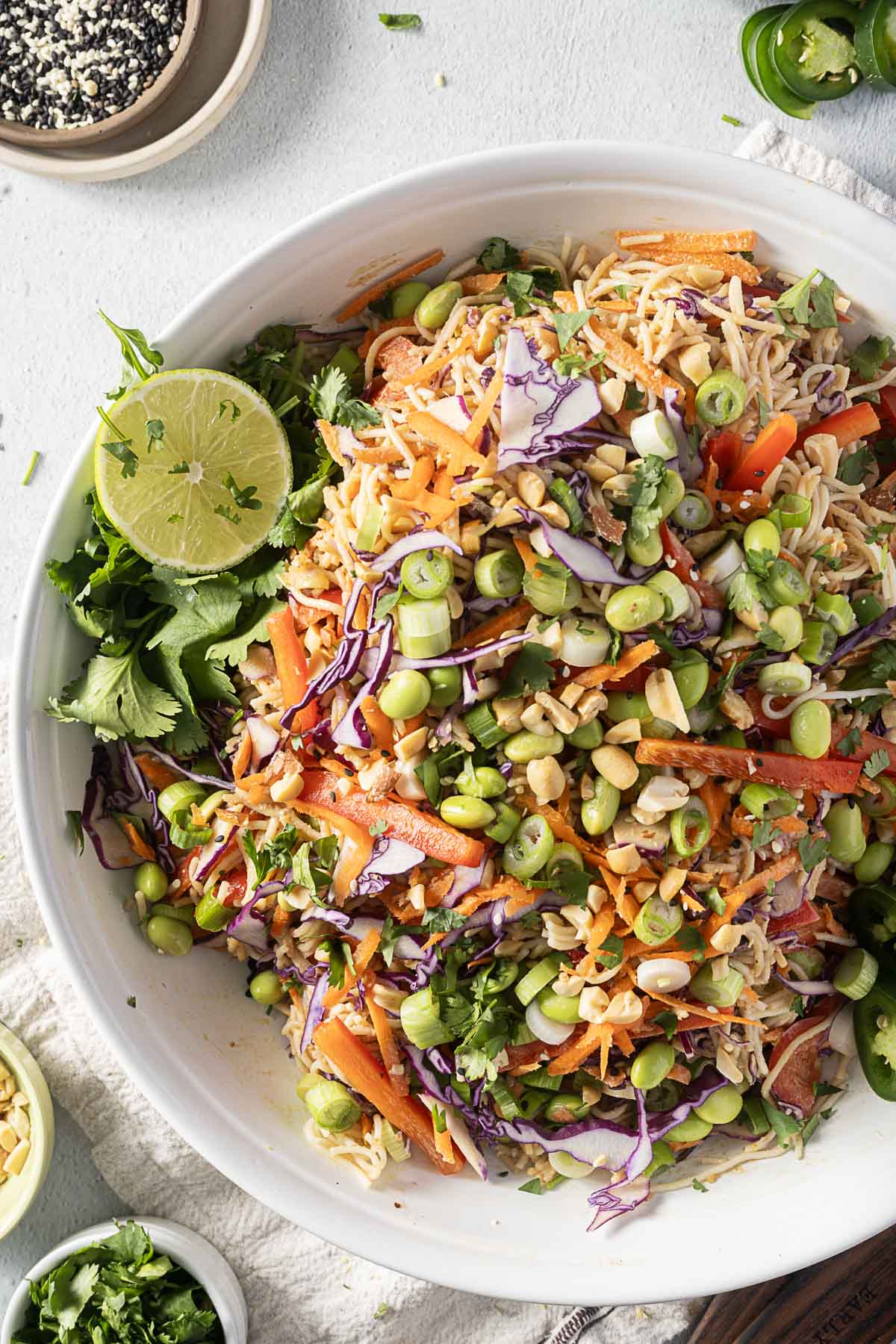 A vibrant cold ramen noodle salad in a white bowl featuring red cabbage, edamame, carrots, peanuts, and cilantro, all enhanced by a drizzle of peanut dressing and garnished with a zesty lime wedge.