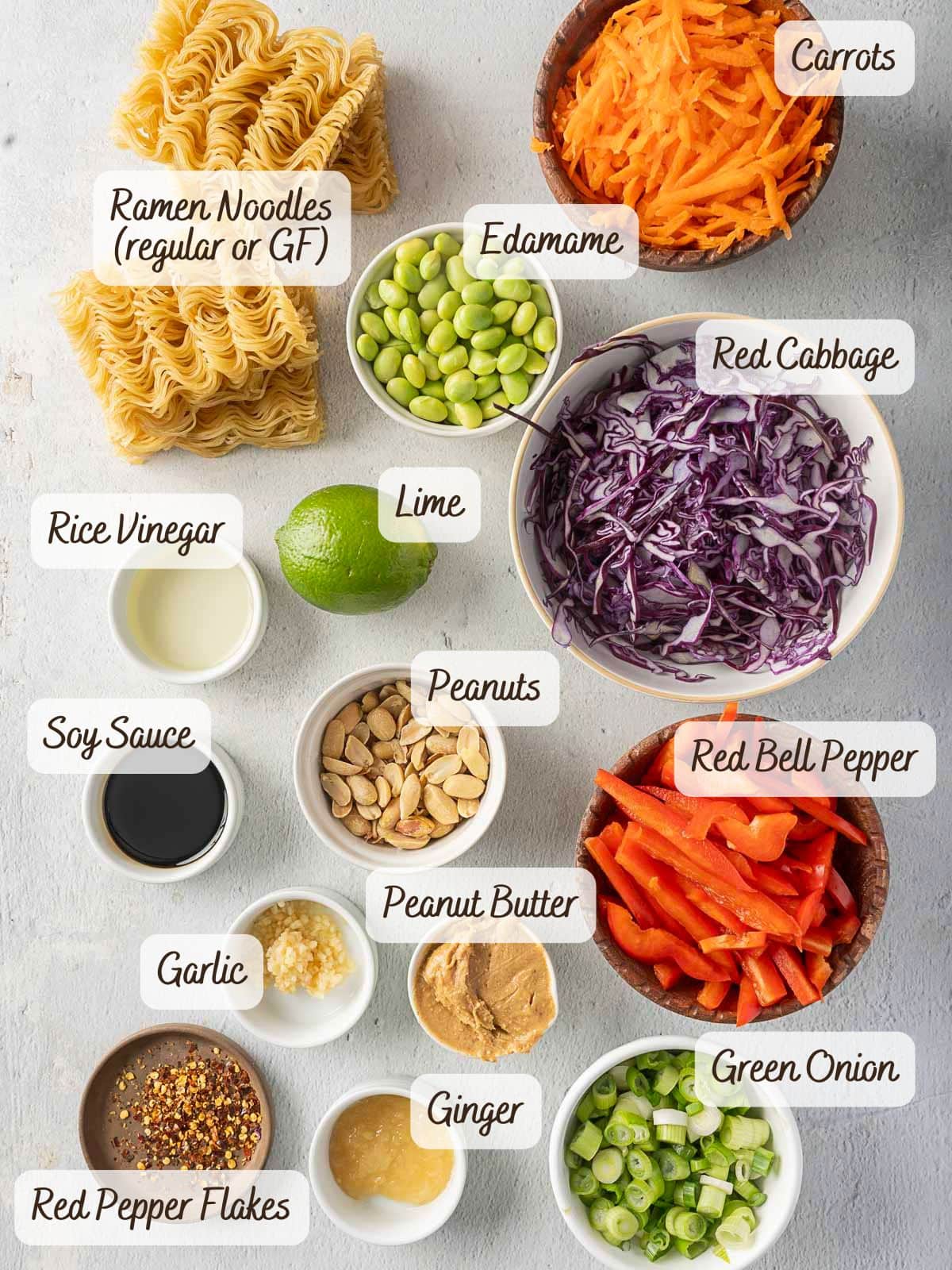 Ingredients for a refreshing cold ramen noodle salad are laid out: ramen noodles (regular or gluten-free), edamame, red cabbage, red bell pepper, carrots, green onion, peanuts, ginger, peanut butter, garlic, soy sauce, rice vinegar, lime.