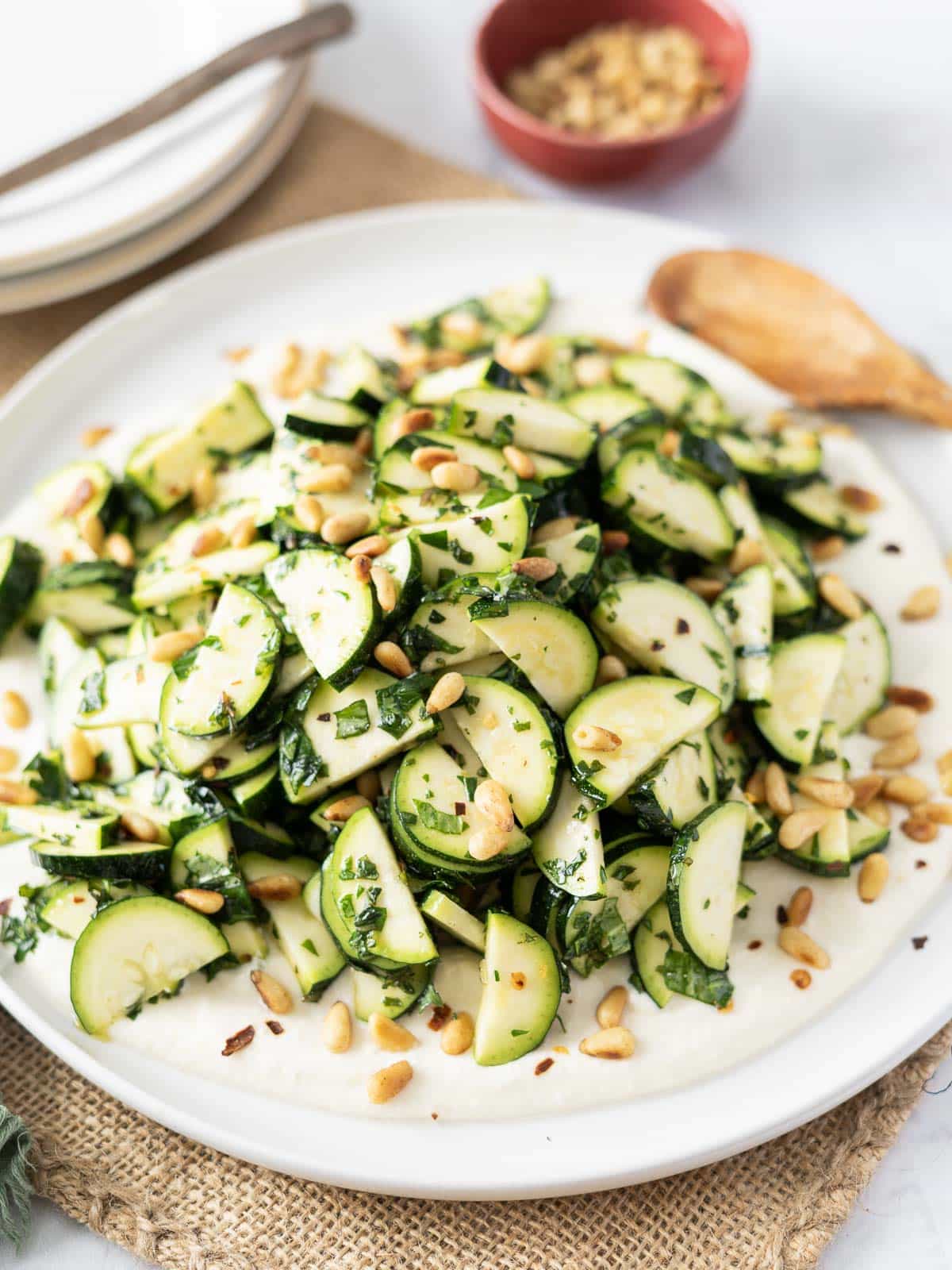 A vibrant zucchini salad featuring sliced zucchini, crunchy pine nuts, and fresh herbs elegantly arranged on a plate, complete with a rustic wooden spoon by its side.