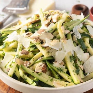 A bowl of zucchini salad with shaved almonds, pecorino cheese, and herbs. A grater and block of cheese are in the background.
