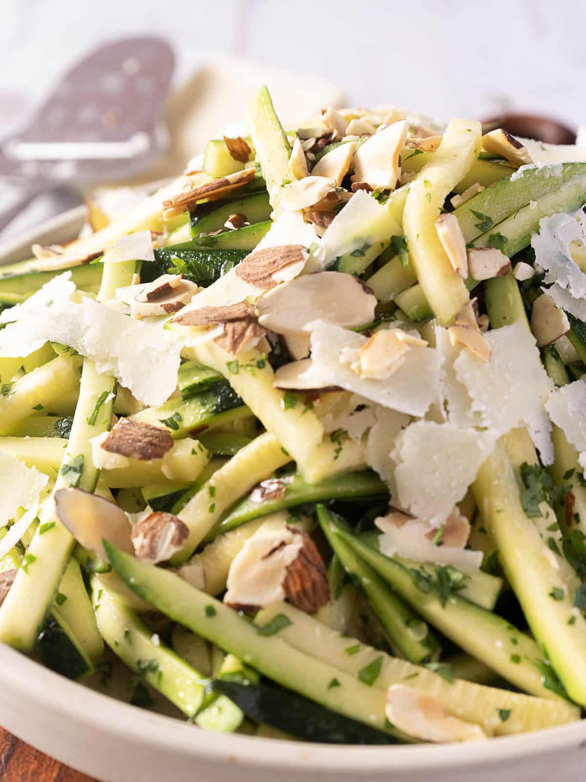 A close-up of a salad with julienned and sautéed zucchini, shaved Pecorino cheese, almond slices, and chopped herbs in a white bowl.