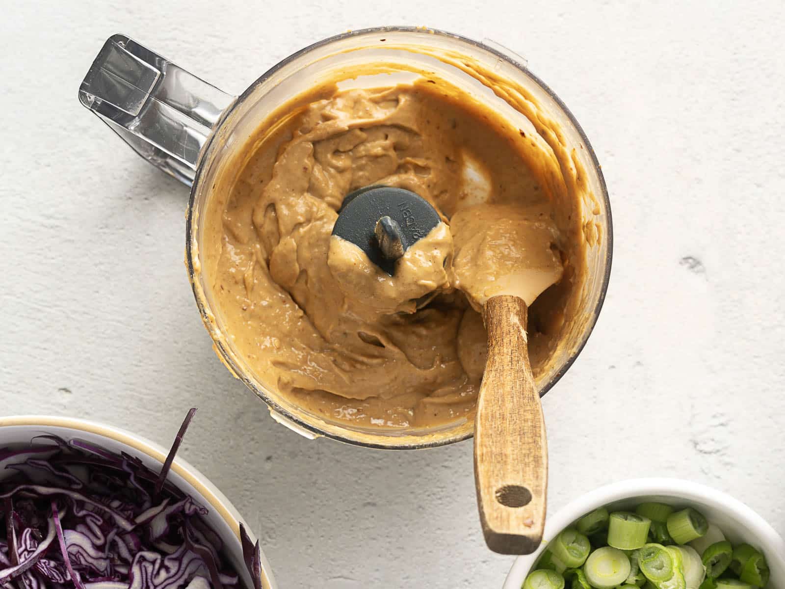 Food processor with creamy beige sauce and spatula. Nearby are bowls of shredded purple cabbage and sliced green onions, perfect for creating a refreshing Cold Ramen Noodle Salad on a light surface.
