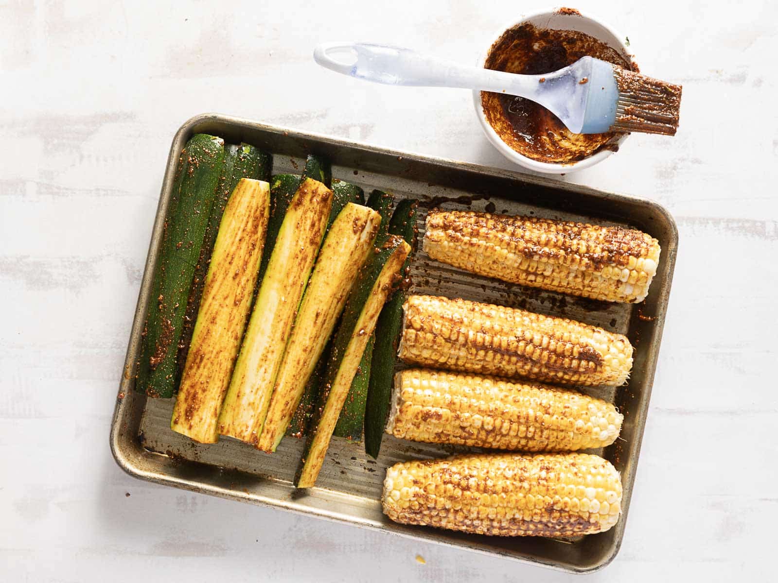 Tray with grilled corn and zucchini spears seasoned to perfection, offering a delightful twist on a classic corn and zucchini salad. Nearby, a brush and bowl brim with tantalizing seasoning sauce.