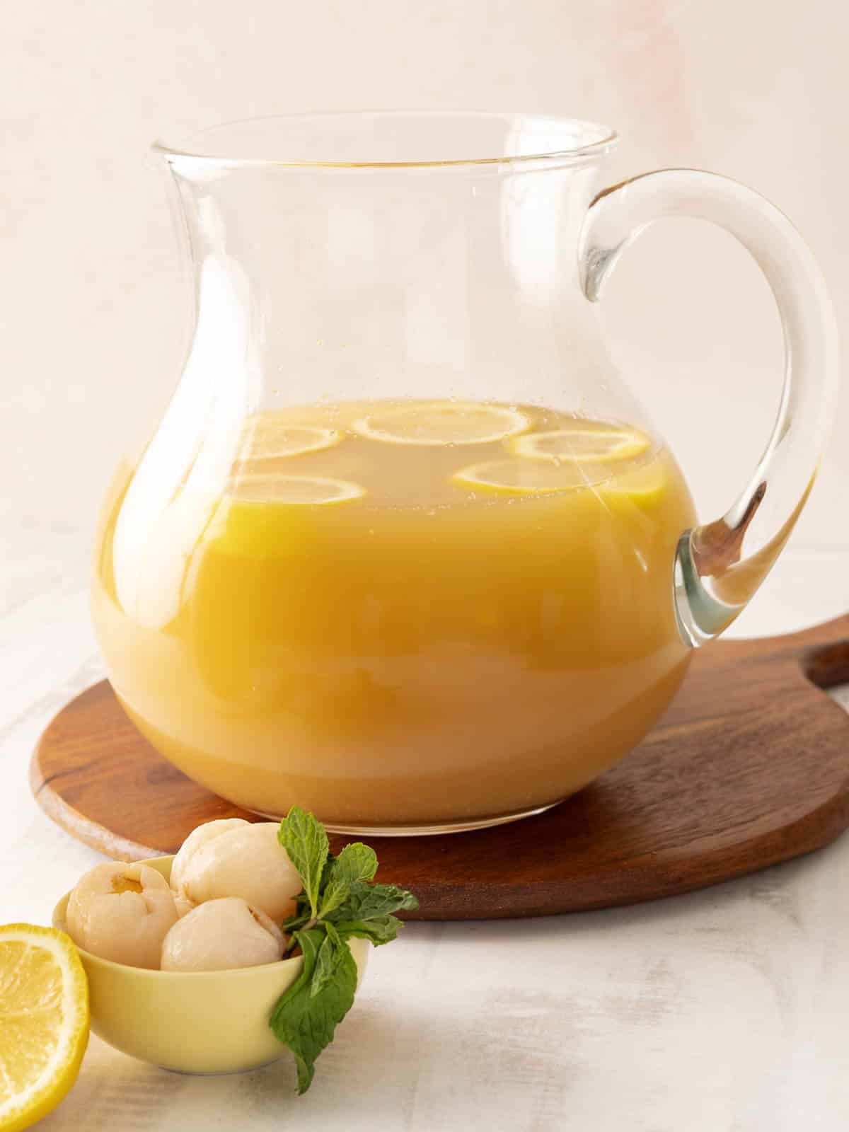 A glass pitcher of refreshing lychee iced tea with lemon slices rests on a wooden board. Nearby, a bowl filled with lychees and a mint garnish adds an extra touch of elegance.