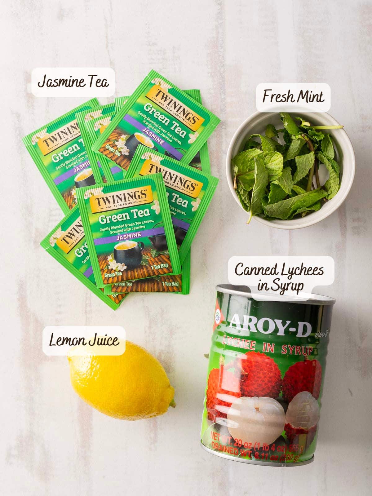 A light surface holds green tea packets, fresh mint, a lemon, and canned lychees with labels, setting the stage for a refreshing Lychee Iced Tea creation.