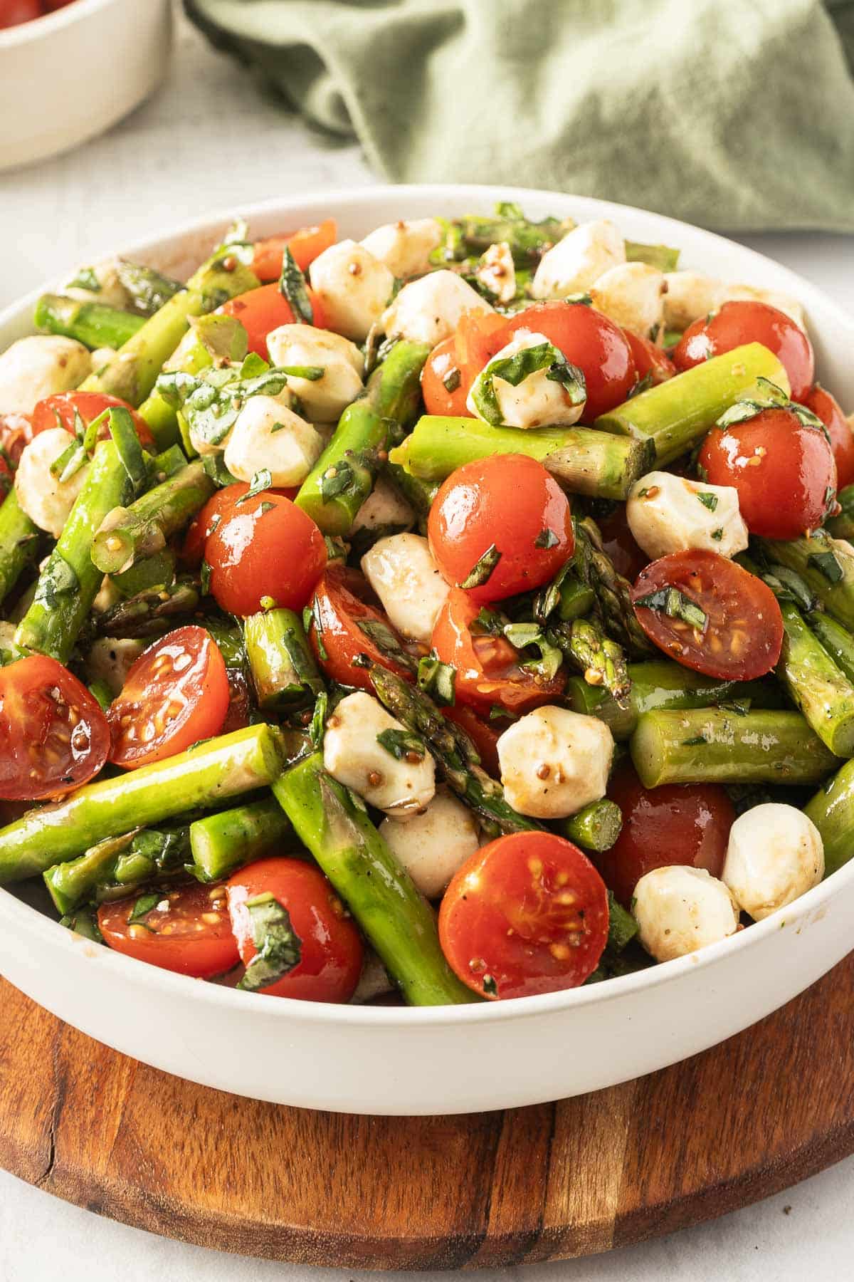 A bowl of grilled asparagus and tomato salad with mozzarella balls rests on a wooden board, offering a vibrant blend of flavors.