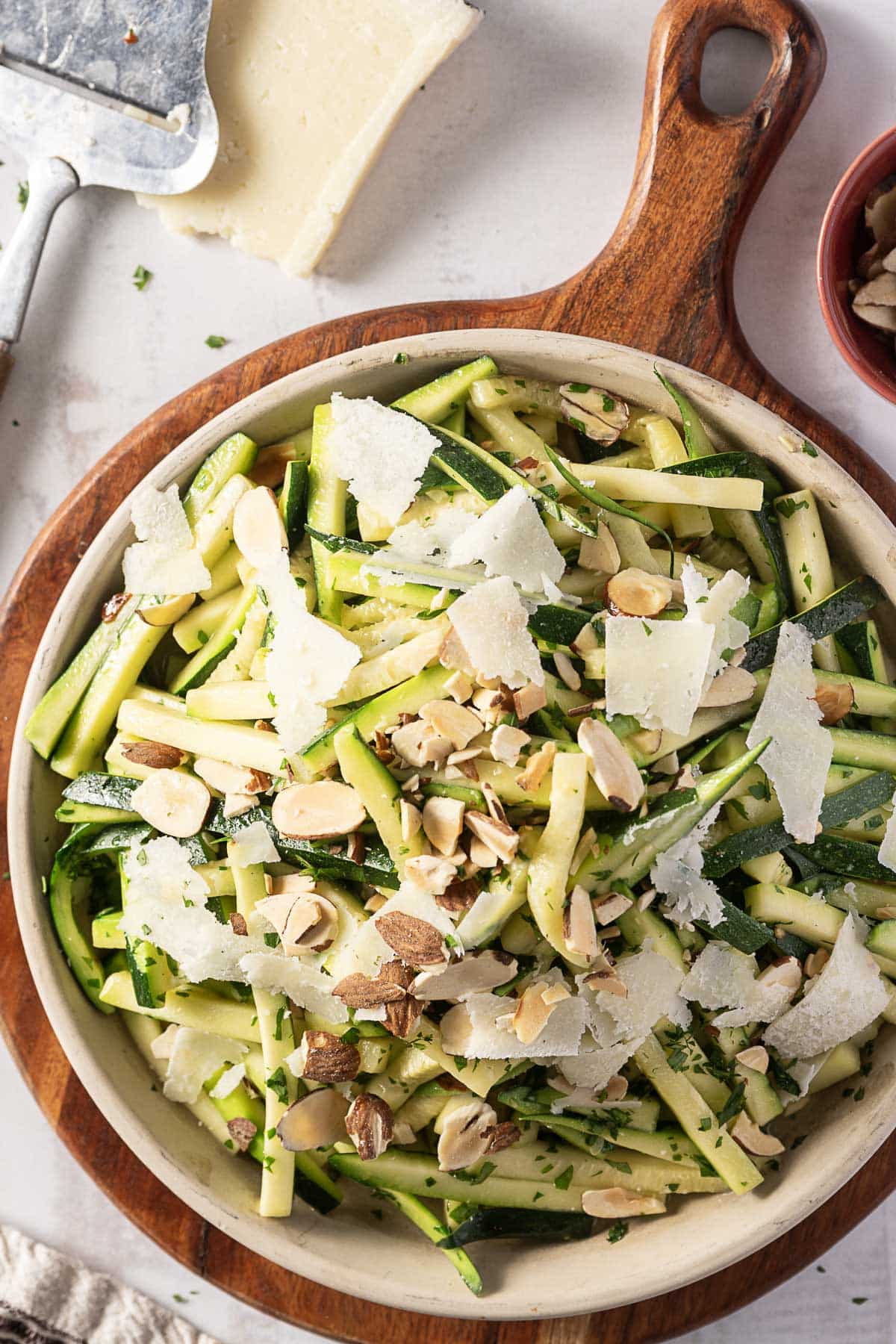 A bowl of zucchini salad with almond slices and Pecorino shavings on a wooden board. Cheese grater and slice of cheese in the background, hinting at the recipe's simplicity and flavor.