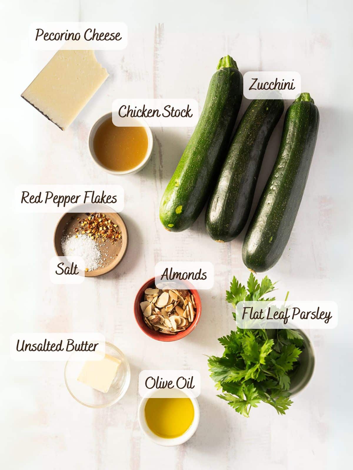 Ingredients on a table: pecorino cheese, chicken stock, sauteed zucchini, red pepper flakes, salt, almonds, flat leaf parsley, unsalted butter, and olive oil.