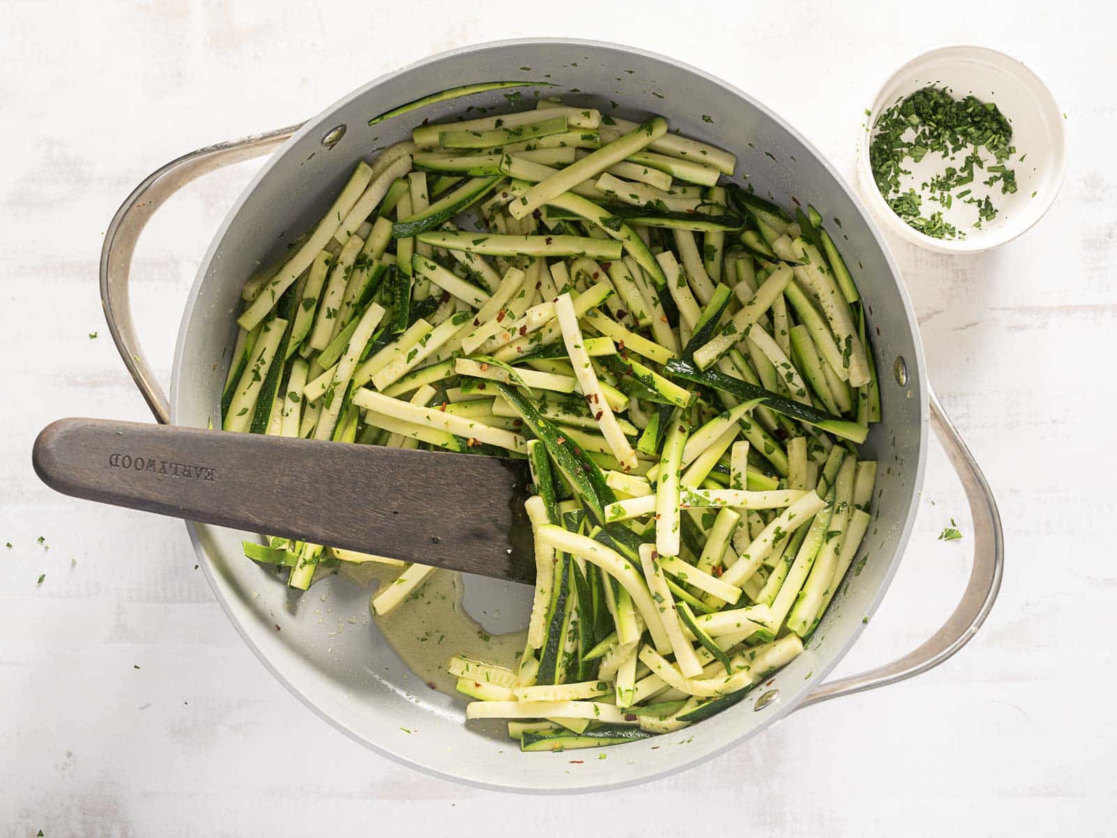 Sliced zucchini being sautéed with a wooden spatula alongside a small bowl of chopped herbs, ready to elevate your favorite recipe. Consider adding a sprinkle of Pecorino for an extra layer of flavor.
