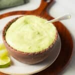 A wooden bowl filled with pale green creamy sauce, with a spoon inside, sits on a plate next to lime wedges.