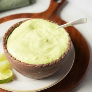 A wooden bowl filled with pale green creamy sauce, with a spoon inside, sits on a plate next to lime wedges.