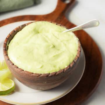 A wooden bowl filled with pale green creamy sauce, with a spoon inside, sits on a plate next to lime wedges.