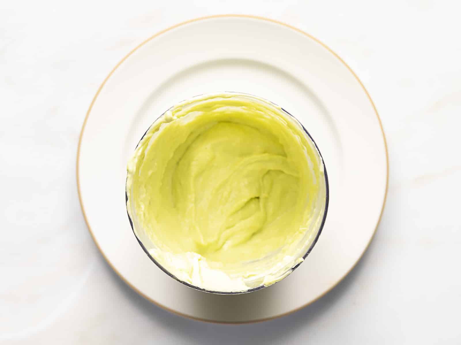 A metal mixing bowl containing smooth, light green avocado cream sits on a white plate with a gold rim, viewed from above on a light surface.