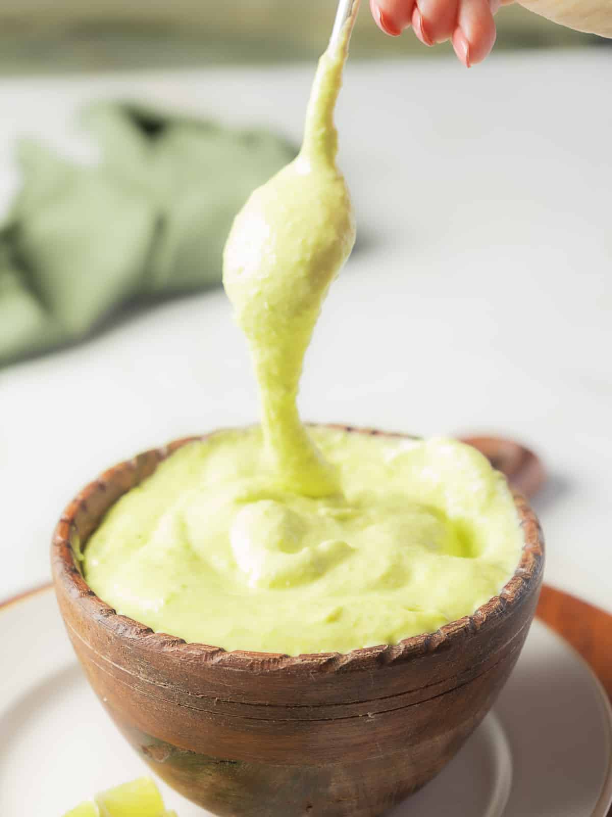 A hand holds a spoonful of thick, creamy green sauce above a wooden bowl filled with the same sauce.