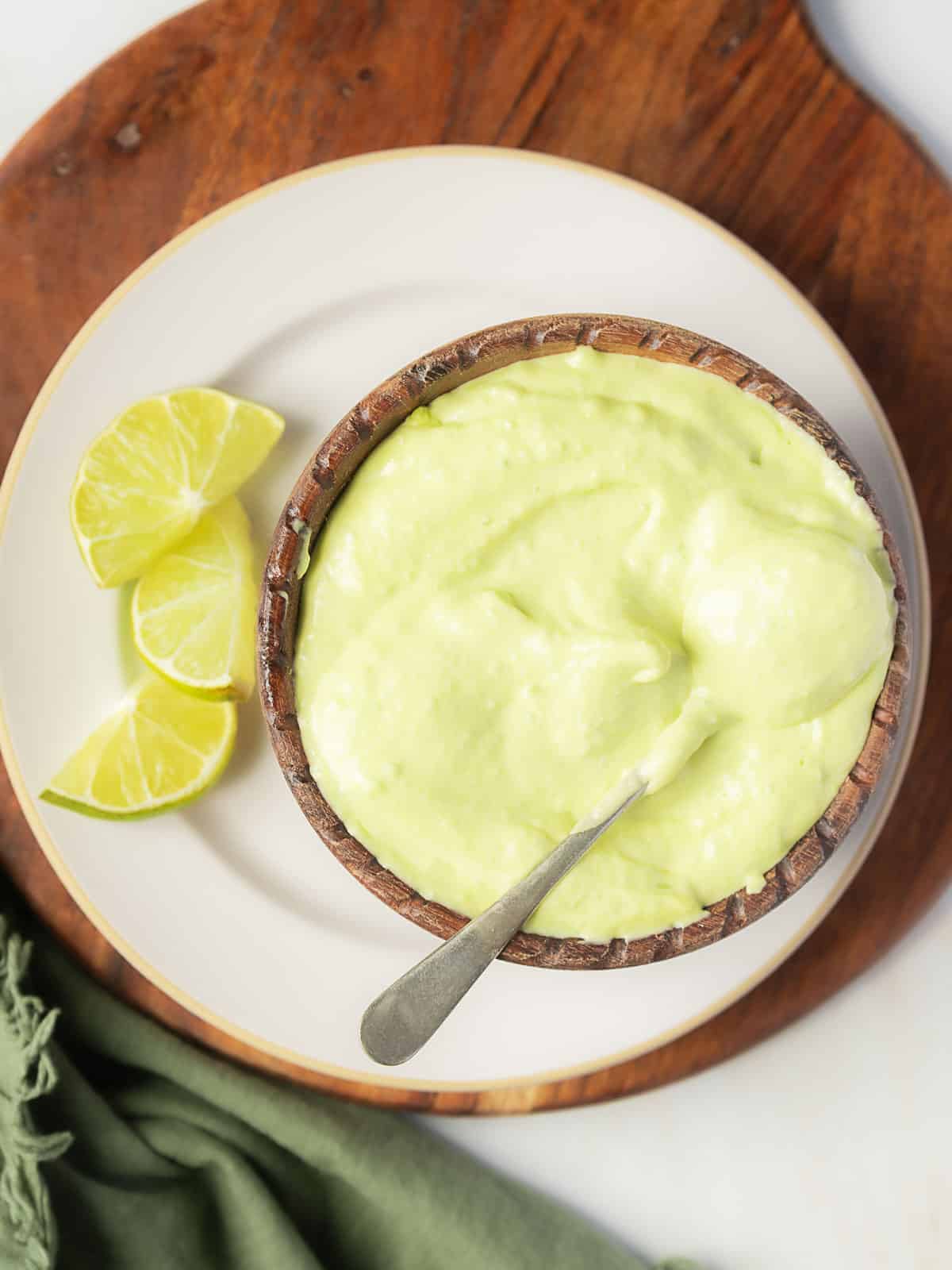 A bowl of creamy green sauce with a spoon, placed on a plate next to three lime wedges on a wooden board.