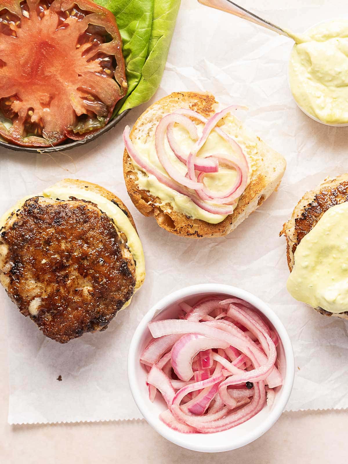 Three burger buns, two with patties and sauce, one with pickled onions, are arranged next to a bowl of pickled onions, a tomato slice, and a lettuce leaf on parchment paper.