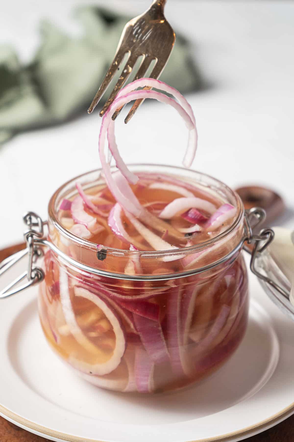 A fork lifts pickled red onion slices from a glass jar filled with brine, placed on a white plate.