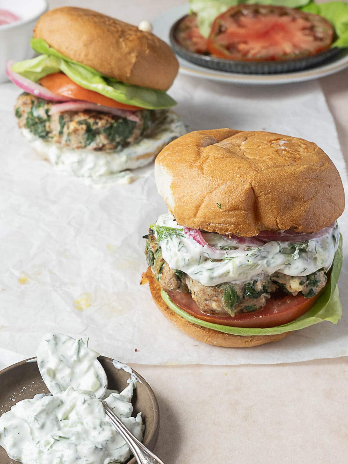Two turkey burgers on buns with lettuce, tomato, red onion, and creamy tzatziki sauce. A plate with sliced tomato and lettuce, and a dish of tzatziki with a spoon are beside the burgers.