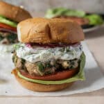 A close-up of a burger with a leafy patty, tomato, lettuce, sliced red onion, and creamy herb sauce on a bun. Another burger sits in the background.