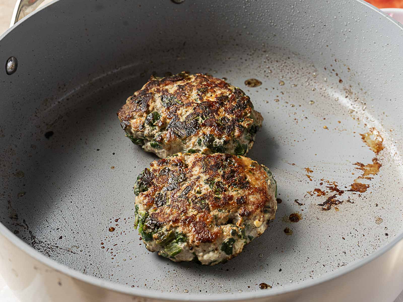 Two cooked burger patties with visible green herbs or vegetables sit in a lightly greased frying pan.