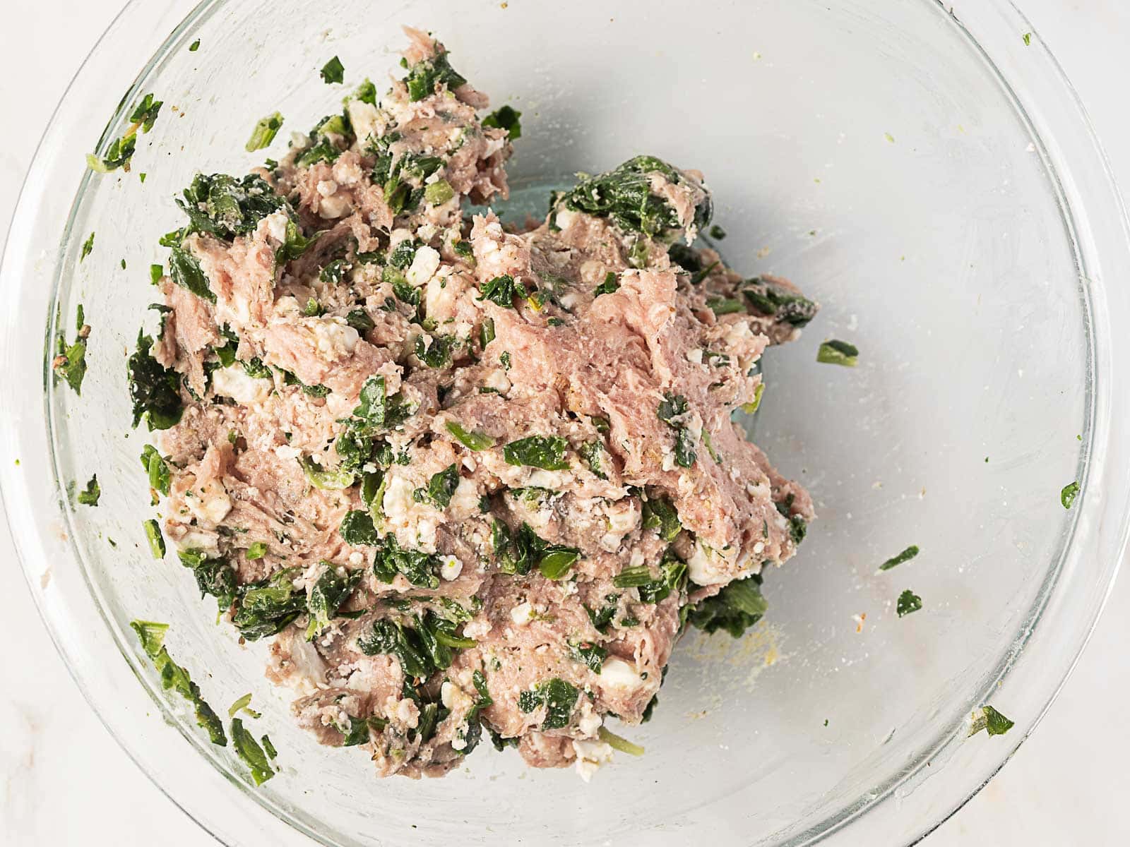 A glass bowl containing a mixture of ground meat, chopped spinach, herbs, and crumbled cheese.