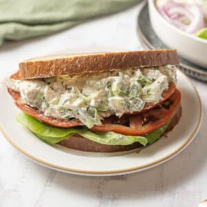 A sandwich with chicken salad, lettuce, tomato, and toasted bread is served on a white plate.