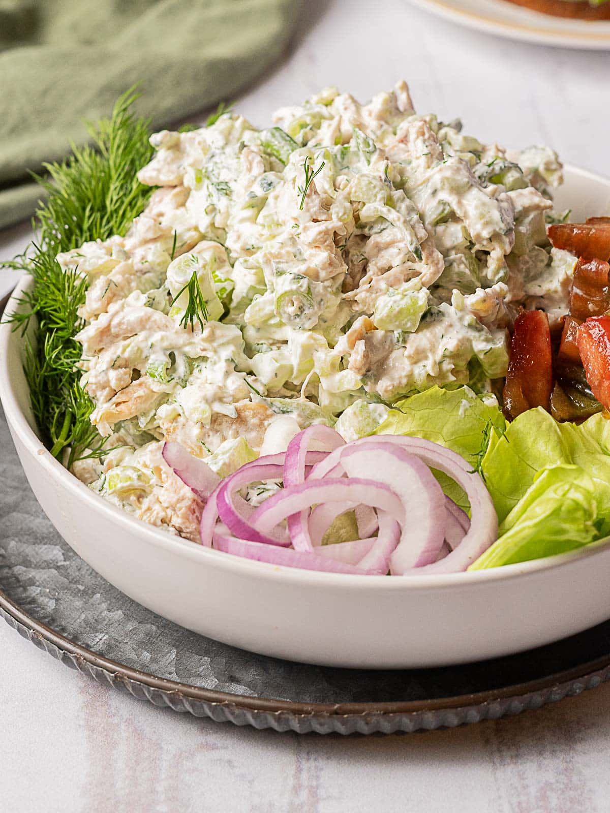 A bowl of tuna salad mixed with mayonnaise and herbs, garnished with fresh dill, served with lettuce, sliced red onion, and tomato wedges.