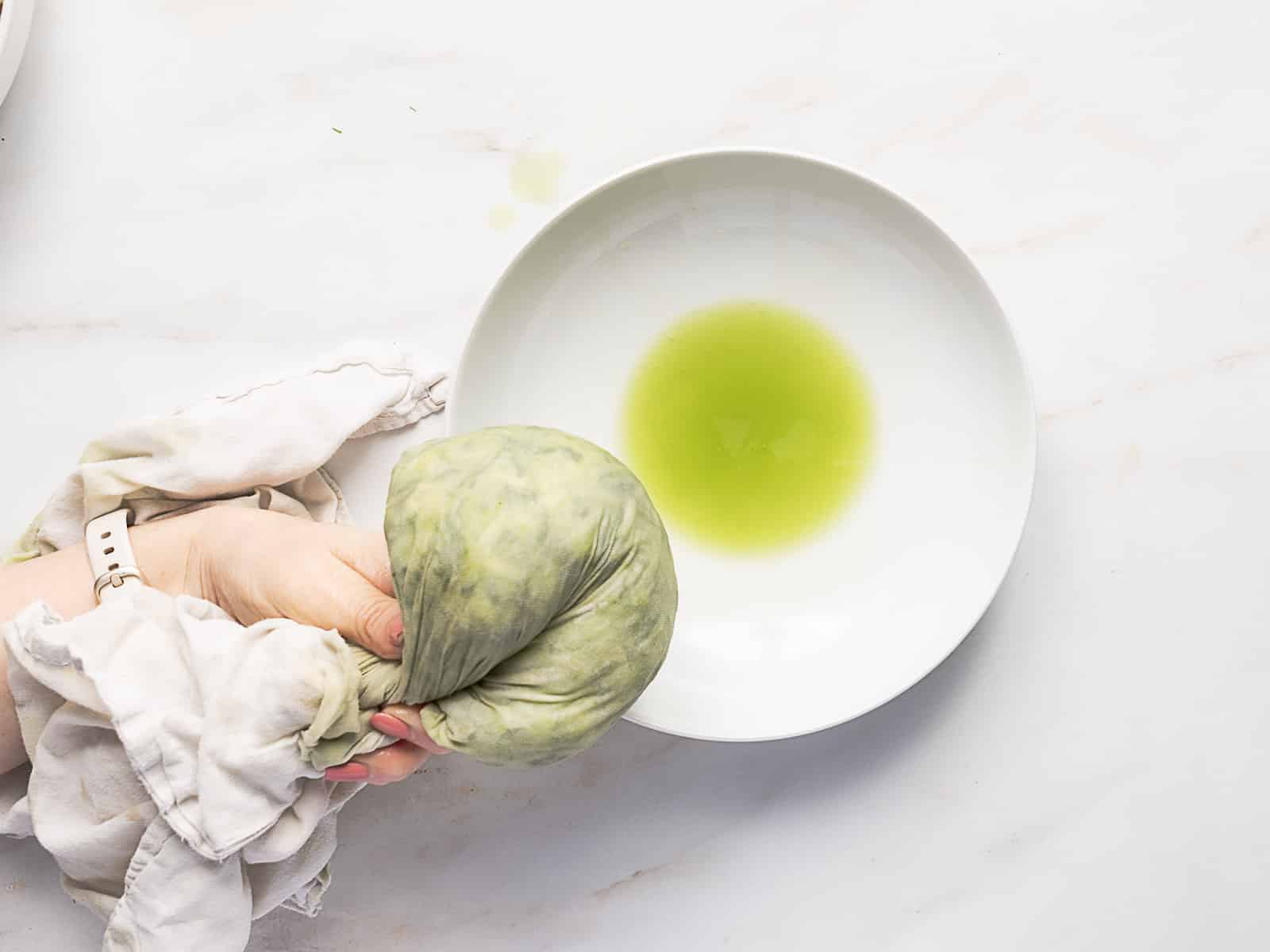 A hand squeezes a cloth bundle of greens over a white bowl, extracting green liquid onto the bowl’s surface.