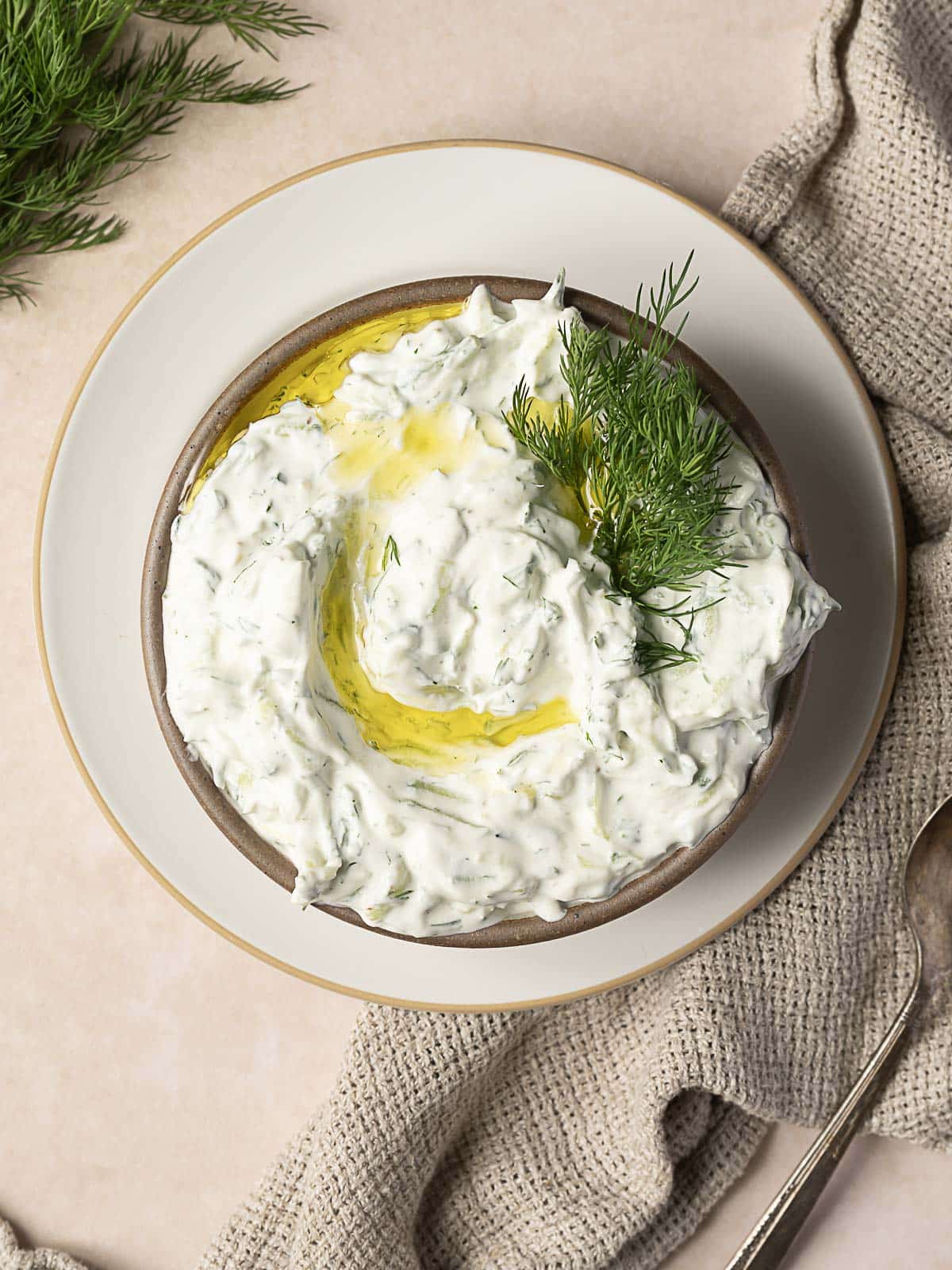 Bowl of creamy tzatziki sauce garnished with fresh dill and a drizzle of olive oil, placed on a white plate with a beige cloth and spoon nearby.