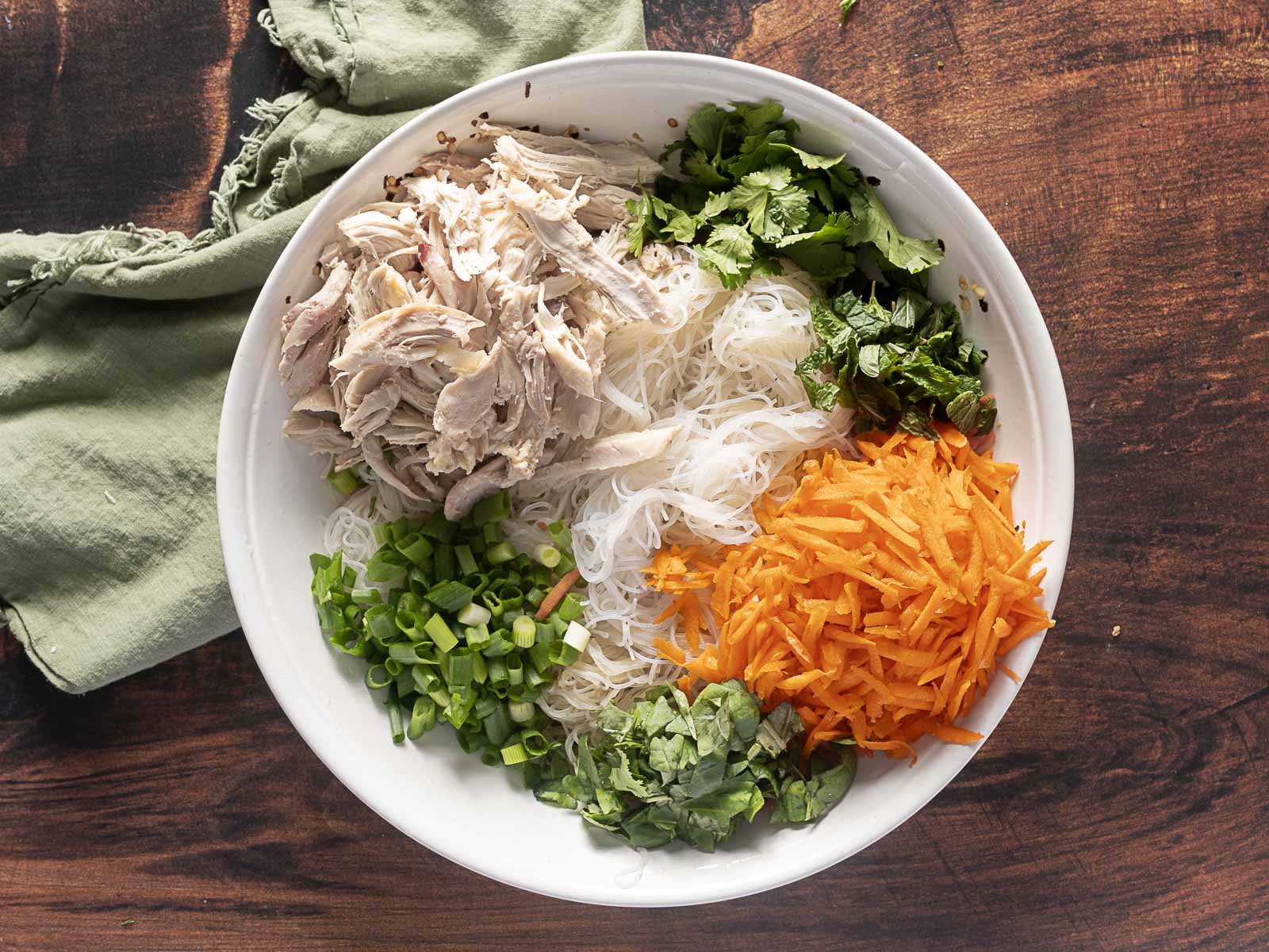 A bowl with shredded chicken, rice noodles, cilantro, mint, grated carrots, green onions, and basil on a wooden surface with a green cloth beside it.