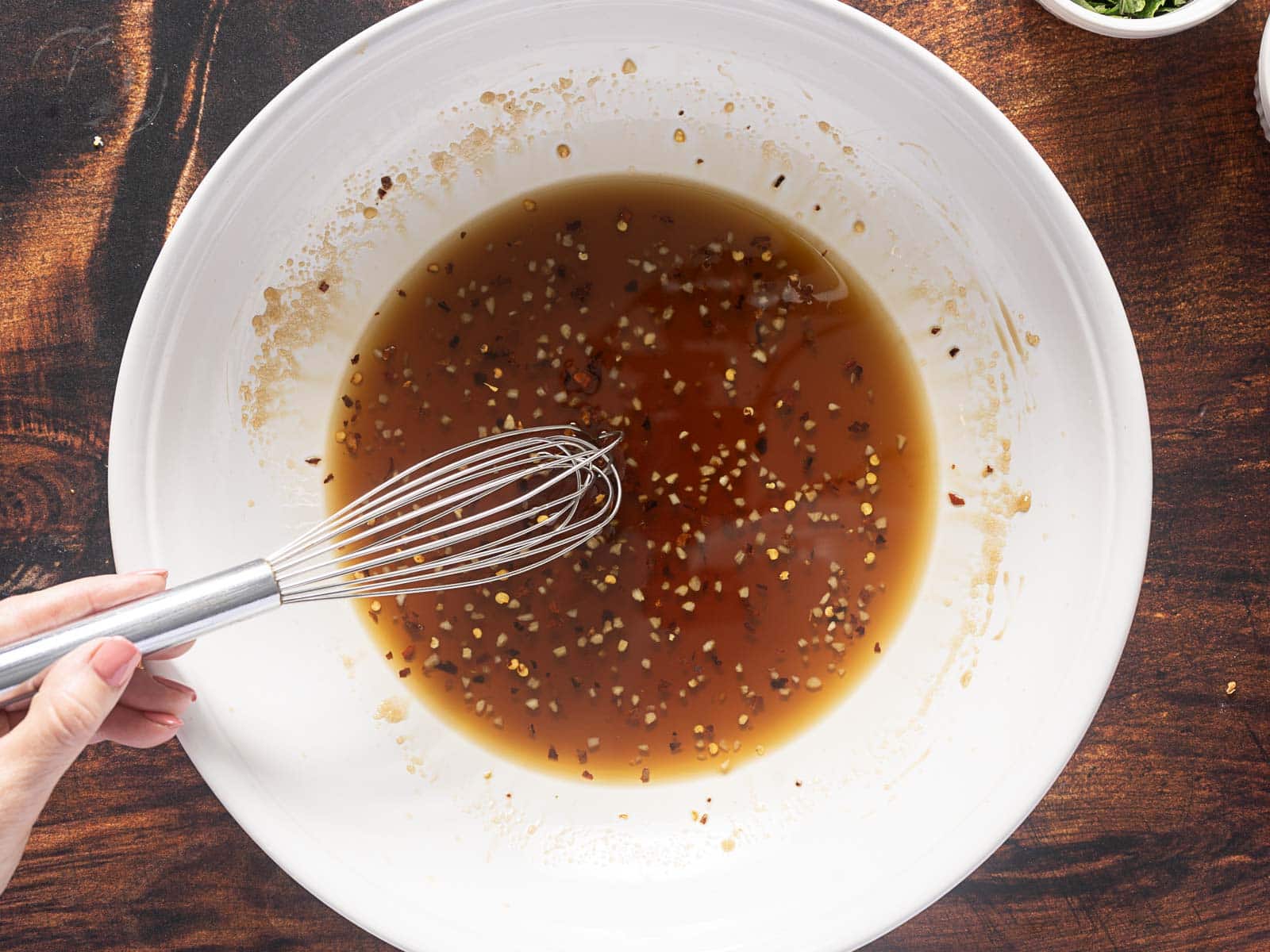 A hand whisking a mixture of dark liquid with seeds in a large white bowl on a wooden surface.