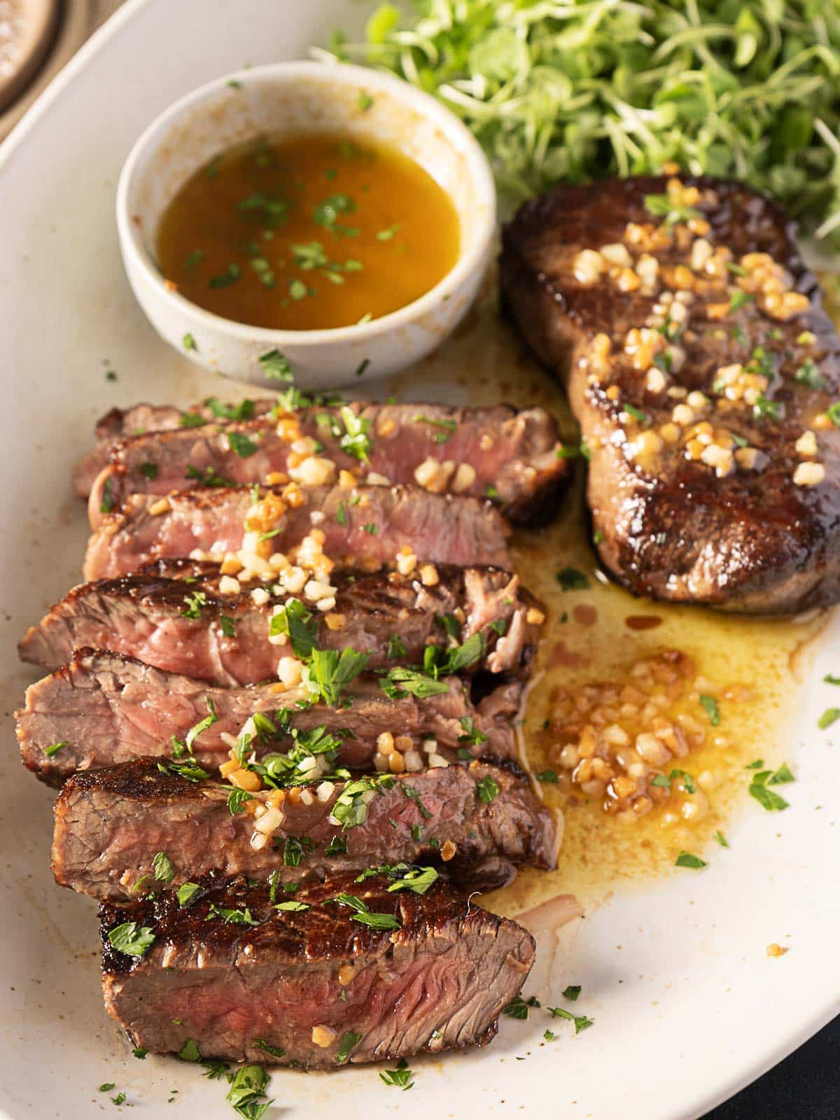 Slices of cooked steak garnished with herbs and garlic are arranged on a plate. A small bowl of sauce and a side of greens accompany the dish.