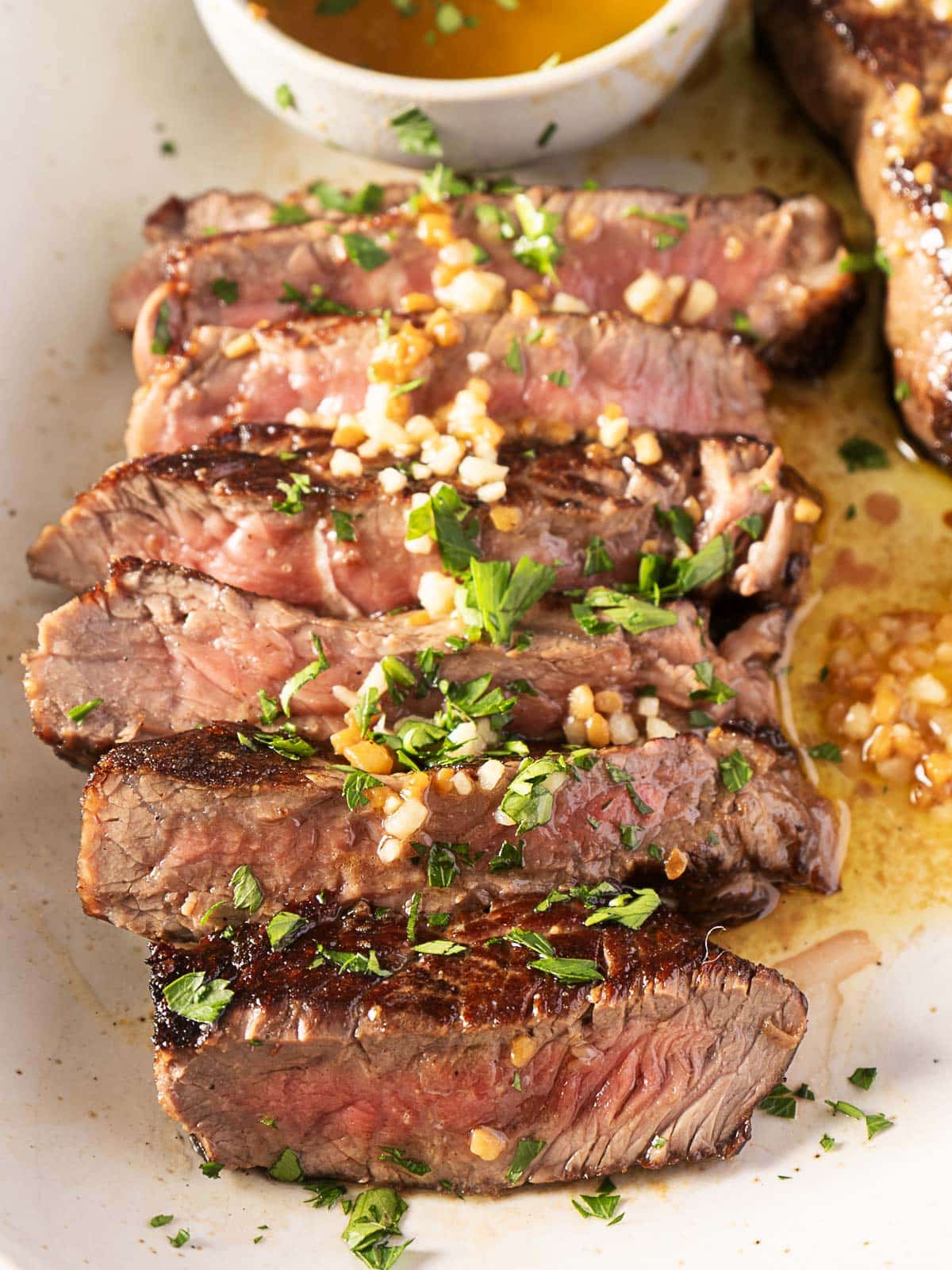 Slices of medium-rare steak topped with chopped garlic and parsley on a white plate, accompanied by a bowl of sauce.