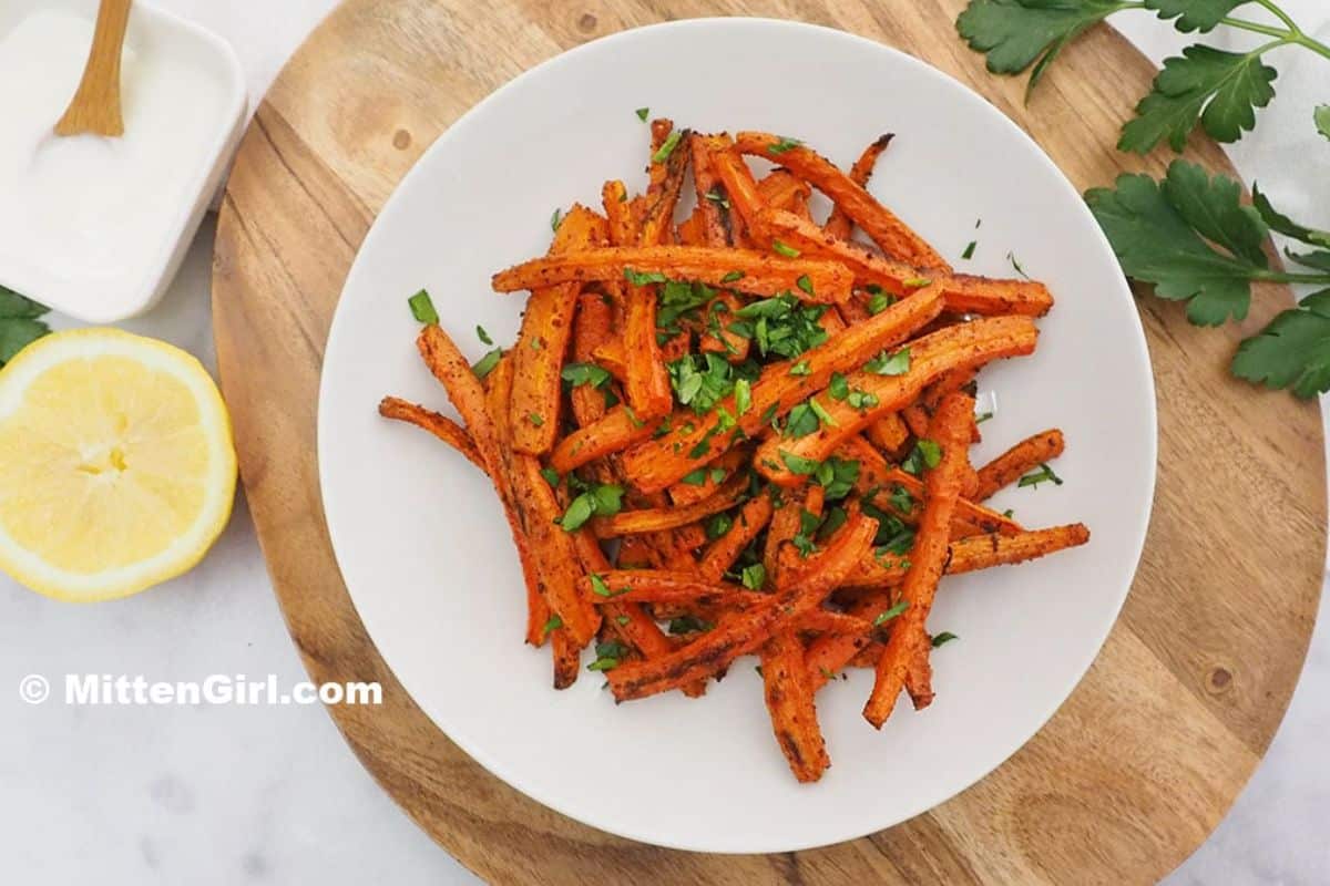 A white plate filled with seasoned carrot fries garnished with chopped herbs showcases the best of fresh carrot recipes. Nearby are a cut lemon, parsley, and a small bowl of white sauce on a wooden cutting board, adding zest to this delightful creation.