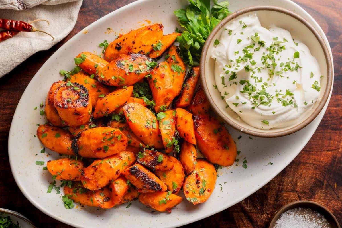 Freshly roasted carrots garnished with herbs grace an oval plate, accompanied by a small bowl of white dip on a wooden table. Perfect for Easter, this delightful dish is a standout in carrot recipes that celebrates the season's finest flavors.