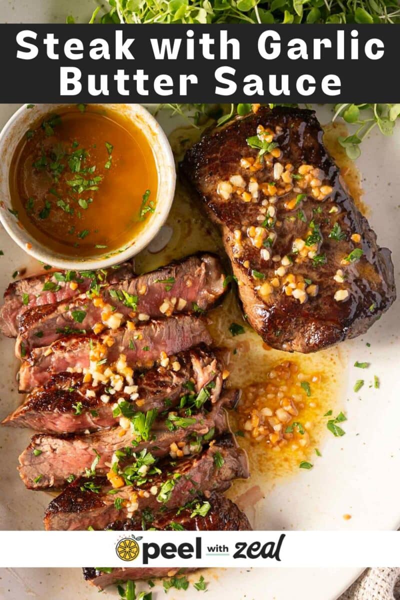 A buttery garlic steak graces the plate, sliced and topped with a rich garlic butter sauce, garnished with herbs beside a small bowl of savory sauce.
