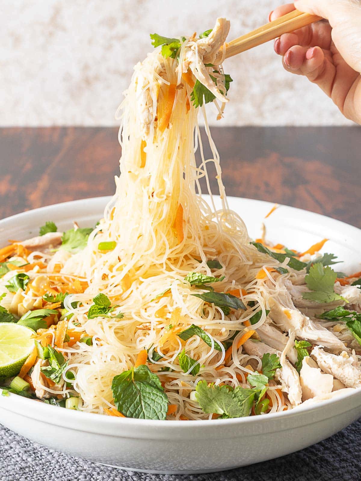 Chopsticks lifting a portion of vermicelli noodles mixed with shredded chicken, carrots, herbs, and lime from a bowl.