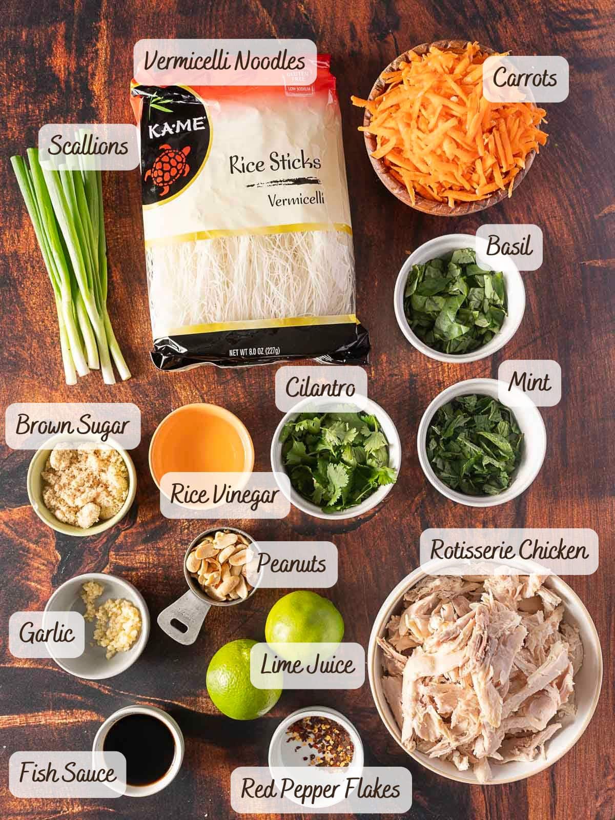 Ingredients for a rice noodle salad are arranged on a wooden surface, featuring vermicelli noodles, carrots, scallions, herbs, brown sugar, vinegar, peanuts, rotisserie chicken, garlic, fish sauce, and lime juice.