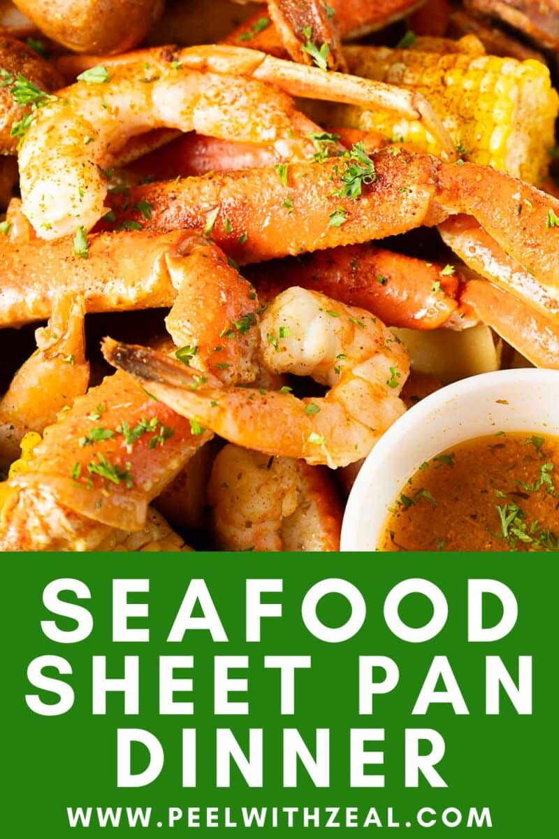 Close-up of assorted cooked seafood bake, including shrimp and crab legs, with corn and potatoes, next to a bowl of dipping sauce.