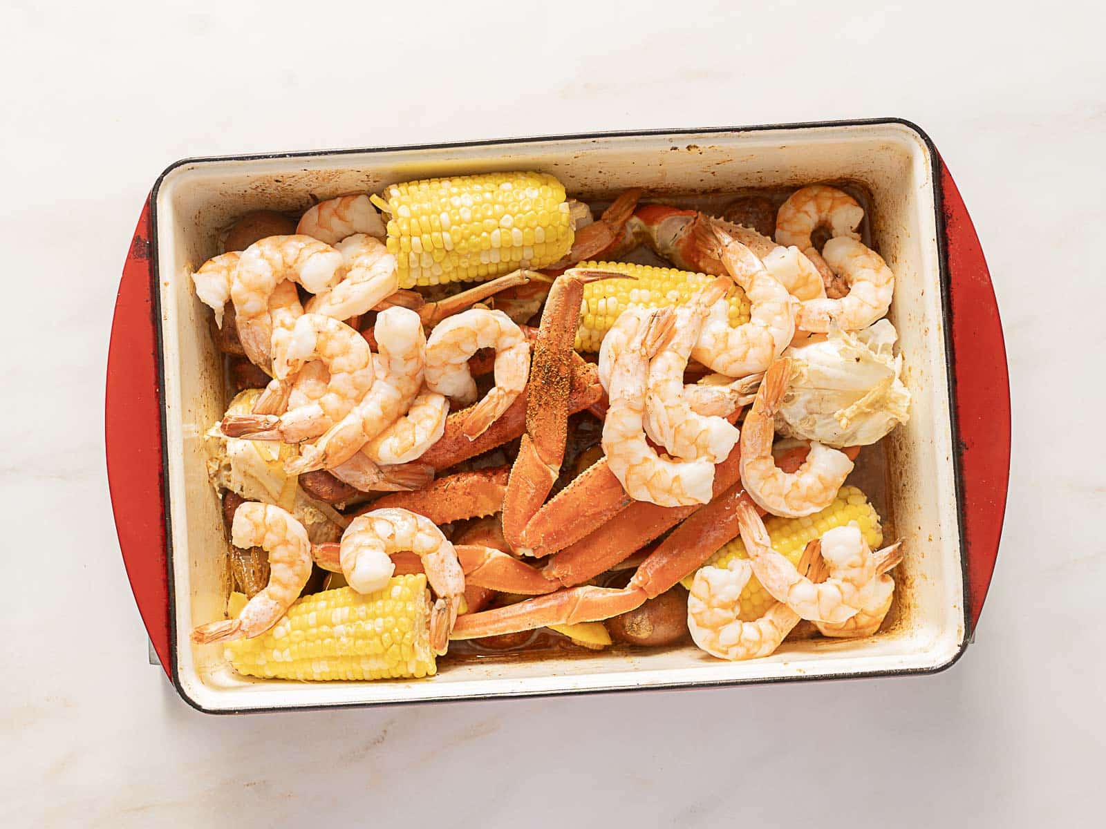 A baking dish filled with cooked shrimp, crab legs, corn on the cob, and sausage pieces on a light surface.