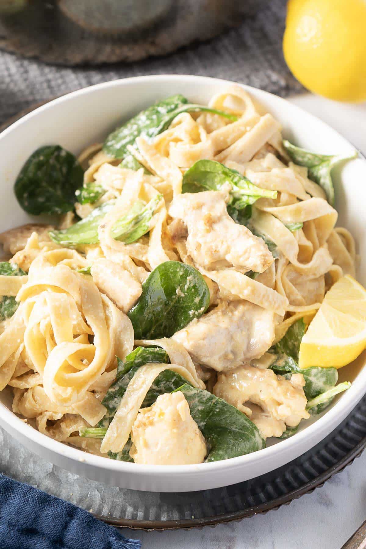 A bowl of creamy pasta with pieces of cooked chicken, spinach, and lemon wedges, placed on a metal tray.