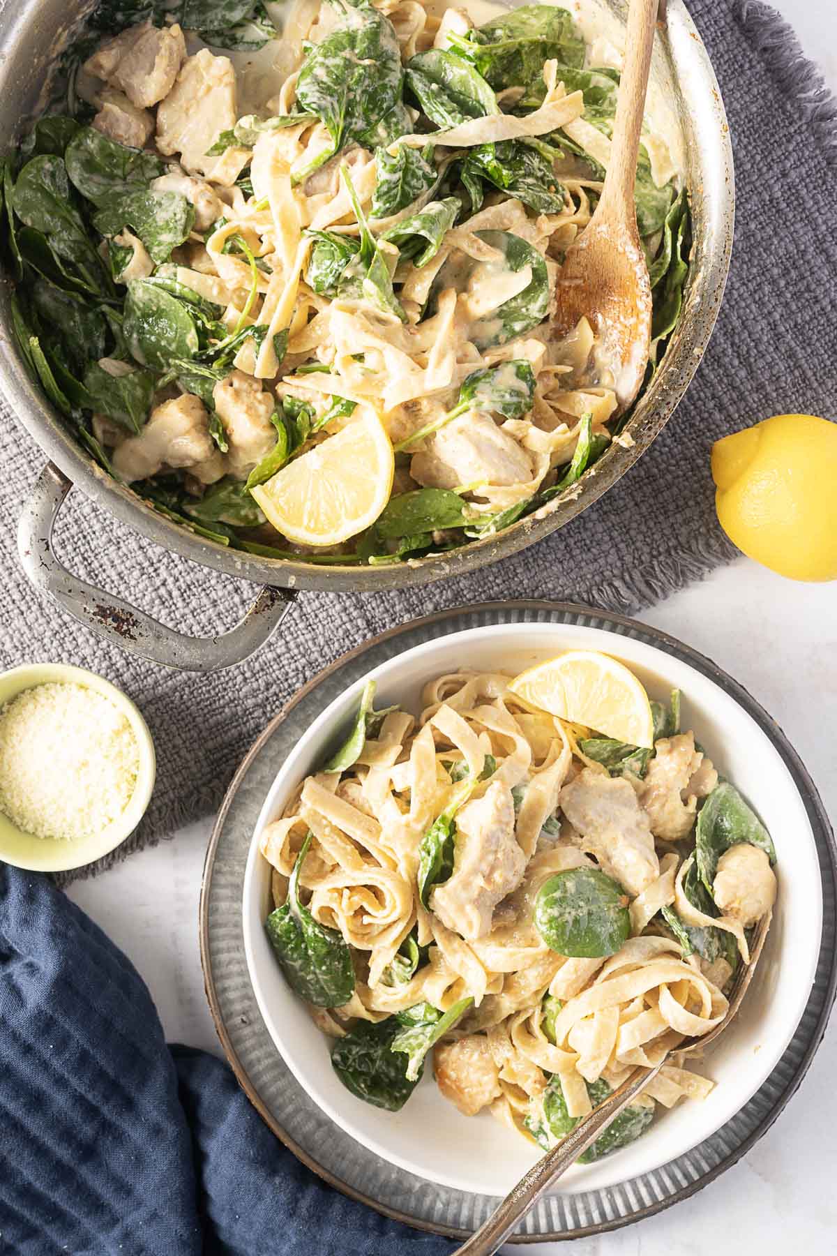A pan and a plate of creamy chicken spinach pasta with lemon, on a gray cloth. A wooden spoon rests in the pan. A lemon and a small bowl of grated cheese are next to them.