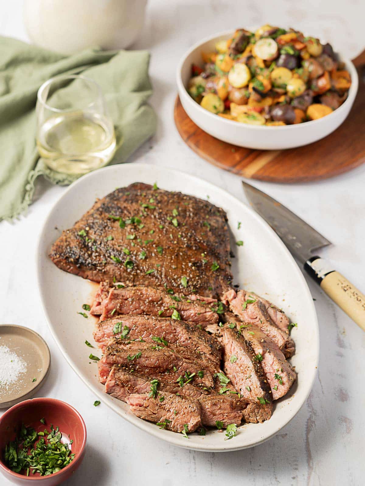 Sliced seasoned steak on a white plate with a knife, glass of white wine, small bowls of salt and herbs, and a dish of roasted vegetables in the background.
