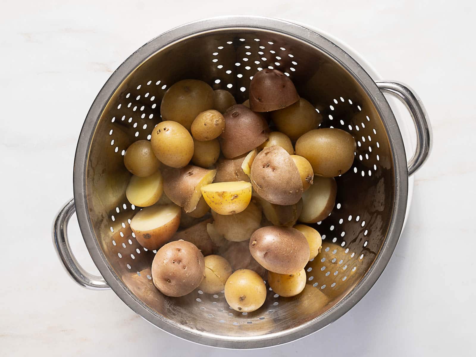 A metal colander filled with washed and halved yellow potatoes on a light surface.