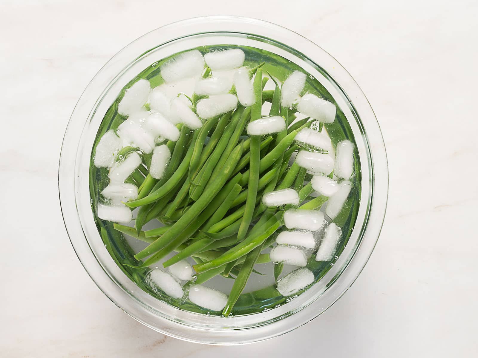 A glass bowl filled with green beans and ice cubes submerged in water, placed on a light-colored surface.