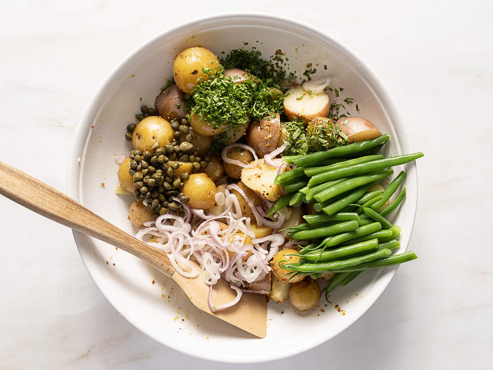 A white bowl contains halved baby potatoes, sliced shallots, capers, chopped herbs, and green beans with a wooden spatula on a white surface.