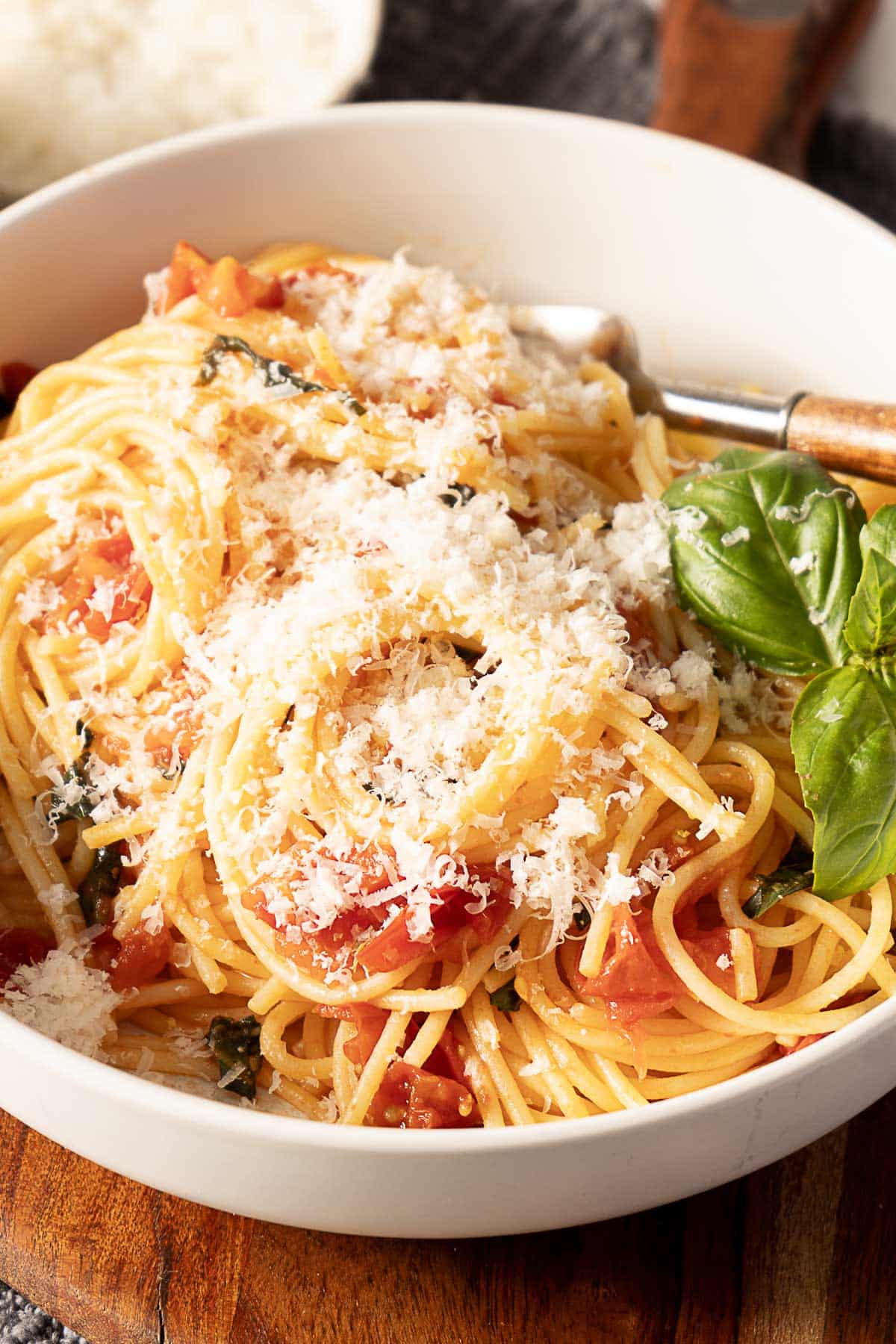 A bowl of spaghetti topped with grated cheese, tomato pieces, and fresh basil leaves, with a fork resting inside the bowl.