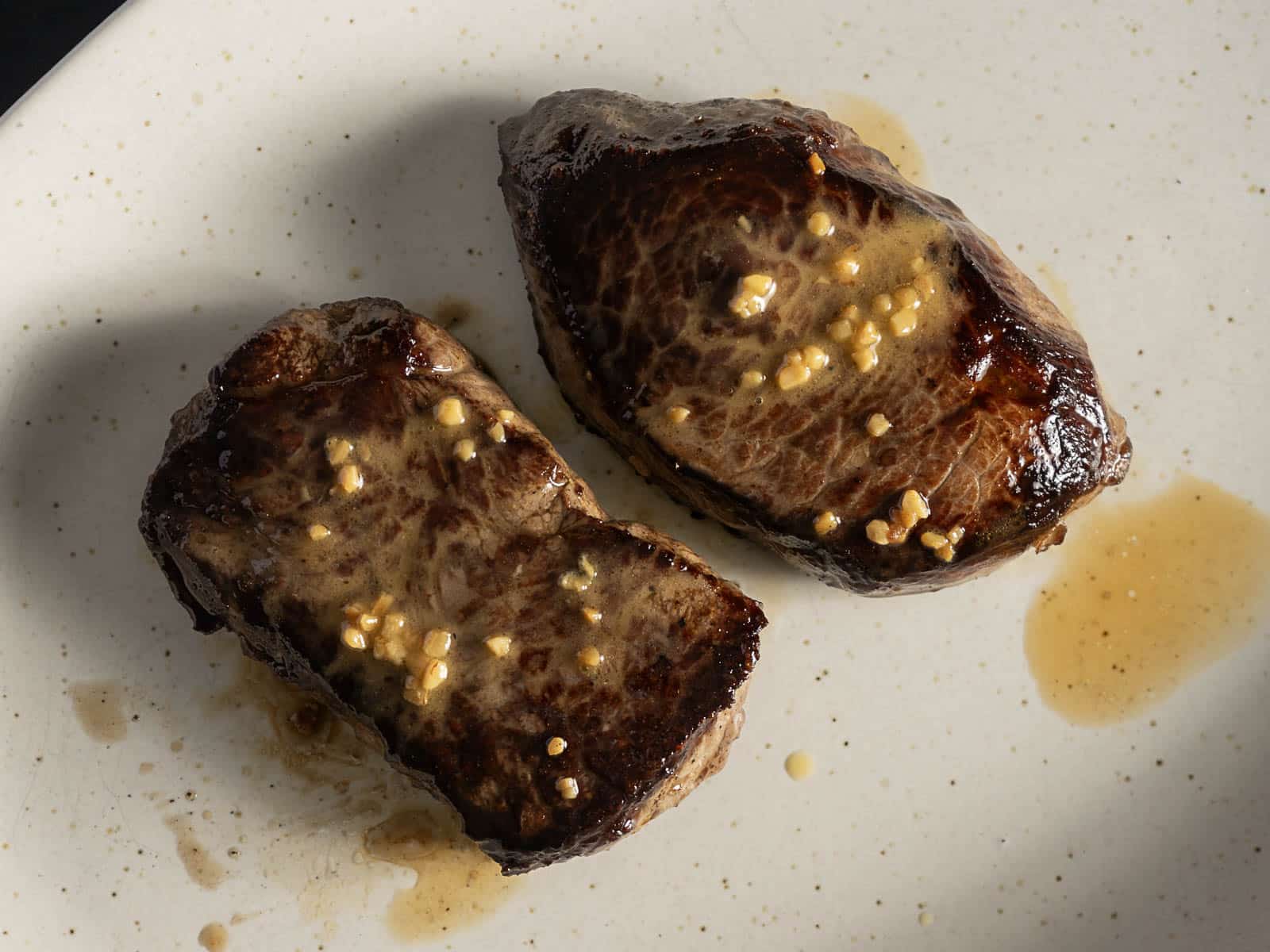 Two pieces of cooked steak with a seared crust and garlic butter sauce on a white plate.